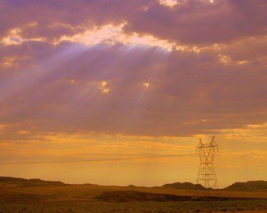 Sunbeams shine through dramatic clouds over a grassy landscape with a distant metal powerline tower. The sky is lit with purple, pink, and orange hues, creating a warm and serene atmosphere.