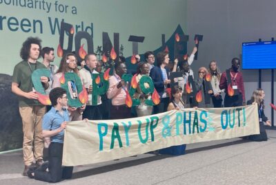 A group of protesters at COP2 in Baku stand in front of a sign saying Pay Up & Phase Out