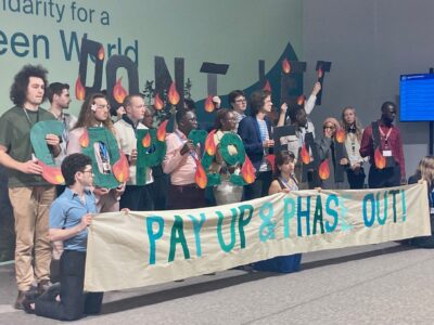 A group of protesters at COP2 in Baku stand in front of a sign saying Pay Up & Phase Out