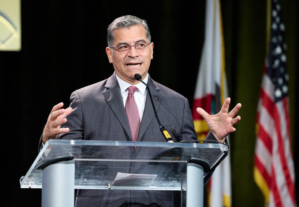 A man in a suit and tie stands at a podium gesturing with his hands, with flags to his side
