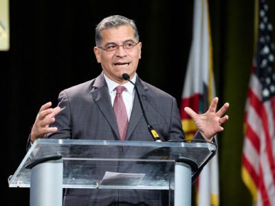 A man in a suit and tie stands at a podium gesturing with his hands, with flags to his side
