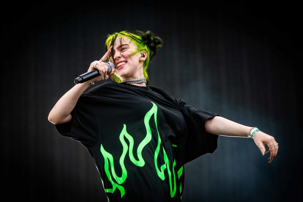 A singer with green hair and a green and black T-shirt performing on stage, smiling with a mic