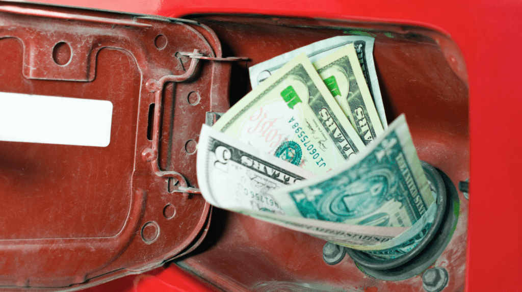 Several US dollar bills are inserted into the fuel tank opening of a red vehicle, with the gas cap door open, symbolizing fuel expenses or the cost of gasoline.