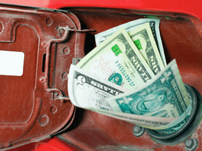 Several US dollar bills are inserted into the fuel tank opening of a red vehicle, with the gas cap door open, symbolizing fuel expenses or the cost of gasoline.