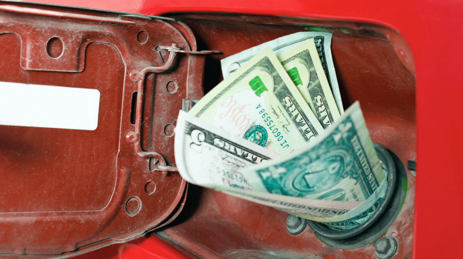 Several US dollar bills are inserted into the fuel tank opening of a red vehicle, with the gas cap door open, symbolizing fuel expenses or the cost of gasoline.