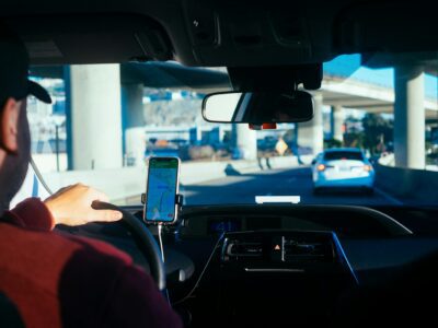 A person drives a car on a highway in daylight, following a white car. A smartphone with a navigation app is mounted on the dashboard. The scene is viewed from the back seat, showing part of the driver’s arm and steering wheel.