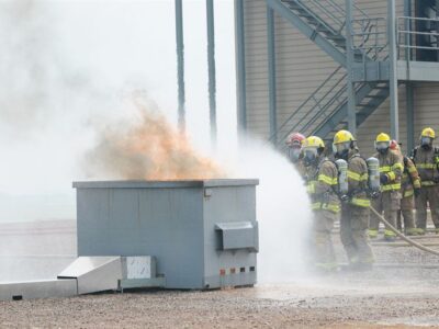 Firefighters in full gear spray water on a flaming dumpster during a training exercise outside, with a building and metal stairs in the background.