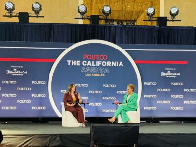 Two women sit on stage having a discussion at the POLITICO The California Agenda event in Los Angeles. The stage has a blue backdrop with event branding and sponsor logos. Both are seated on white chairs.
