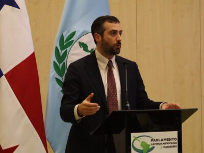 A man in a suit and tie speaks at a podium with Parlamento Latinoamericano y Caribeño signage, standing between the flags of Panama and another country.