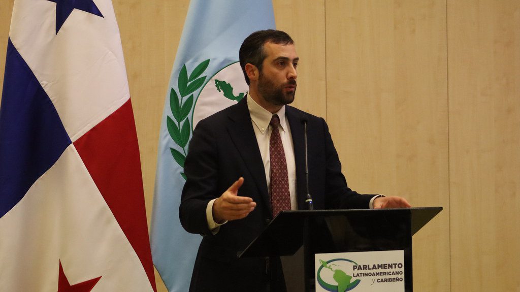 A man in a suit and tie speaks at a podium with Parlamento Latinoamericano y Caribeño signage, standing between the flags of Panama and another country.