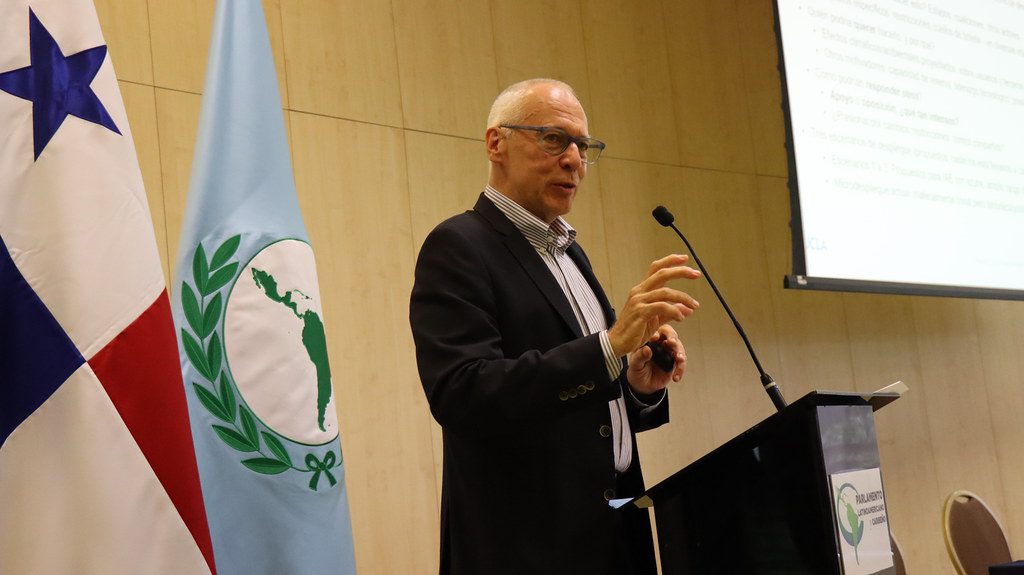 A man in a suit speaks at a podium with a microphone. Behind him are a flag with a blue star and a light blue flag with a green laurel and map. A screen displaying text is visible in the background.