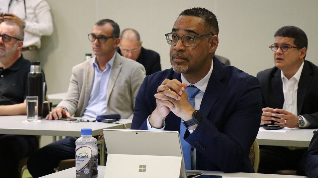 A man in a suit and blue tie sits at a desk with a laptop and water bottle, attentively listening in a meeting room with other seated people in the background.