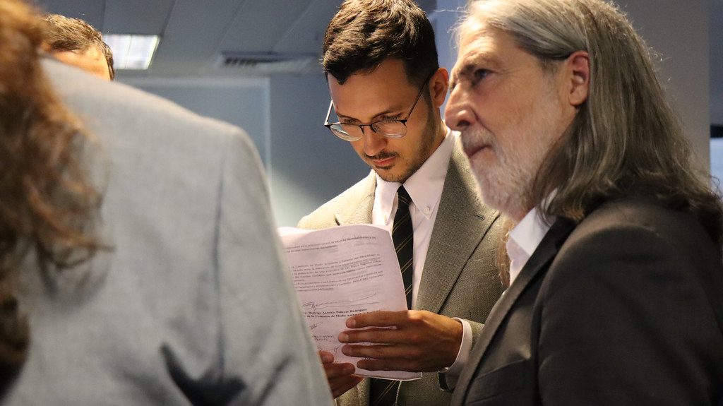 Two men in suits stand indoors; one with long gray hair looks ahead while the other, with glasses, examines documents. Office ceiling panels and fluorescent lights are in the background.