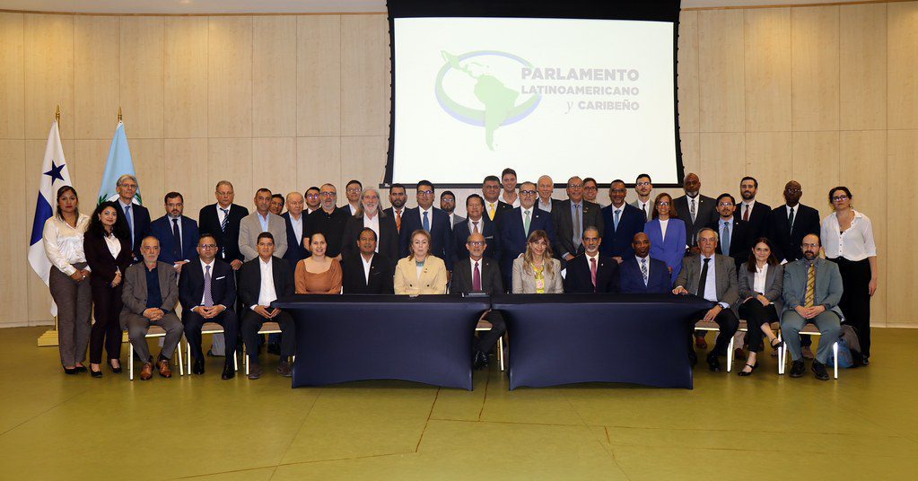 A large group of men and women in business attire pose for a formal photo in front of a Parlamento Latinoamericano y Caribeño logo, with two flags on the left and three people seated at a table in the center.