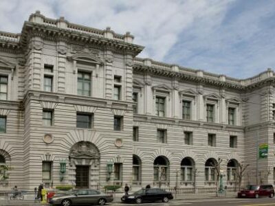 A large, historic building with ornate stone detailing, arched windows on the ground floor, and columns between upper windows. Cars and people are visible on the street in front. The sky is partly cloudy.