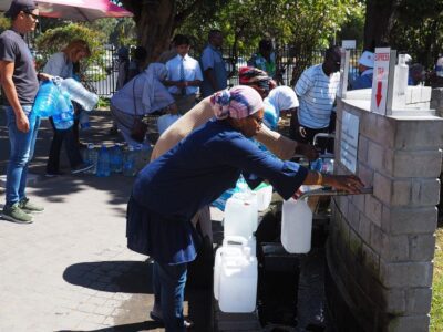 People fill water containers at an outdoor water collection point, with several waiting and others carrying filled bottles. Sunlight casts shadows, and trees provide partial shade in the background.