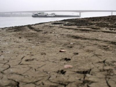 A cracked, dry riverbed in the foreground with a large river and a bridge in the background; a boat is visible on the water under a gray, overcast sky.