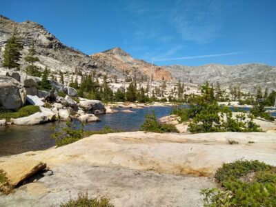 A clear mountain lake surrounded by rocky terrain, pine trees, and rugged peaks under a bright blue sky with scattered clouds.