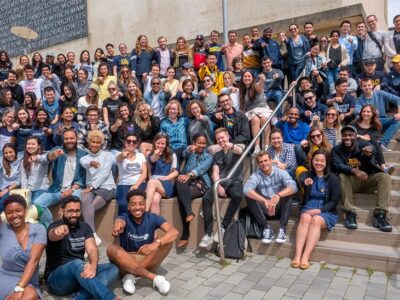 A large, diverse group of people sits and stands on outdoor steps, smiling and posing for a group photo in front of a building on a sunny day.