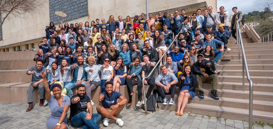 A large, diverse group of people sits and stands on outdoor steps, smiling and posing for a group photo in front of a building on a sunny day.