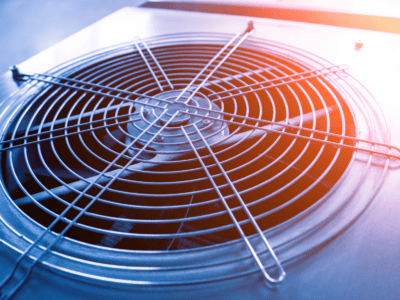 Close-up of a metal ventilation fan grille on an industrial air conditioning or cooling unit, with sunlight creating a blue and orange gradient across the surface.