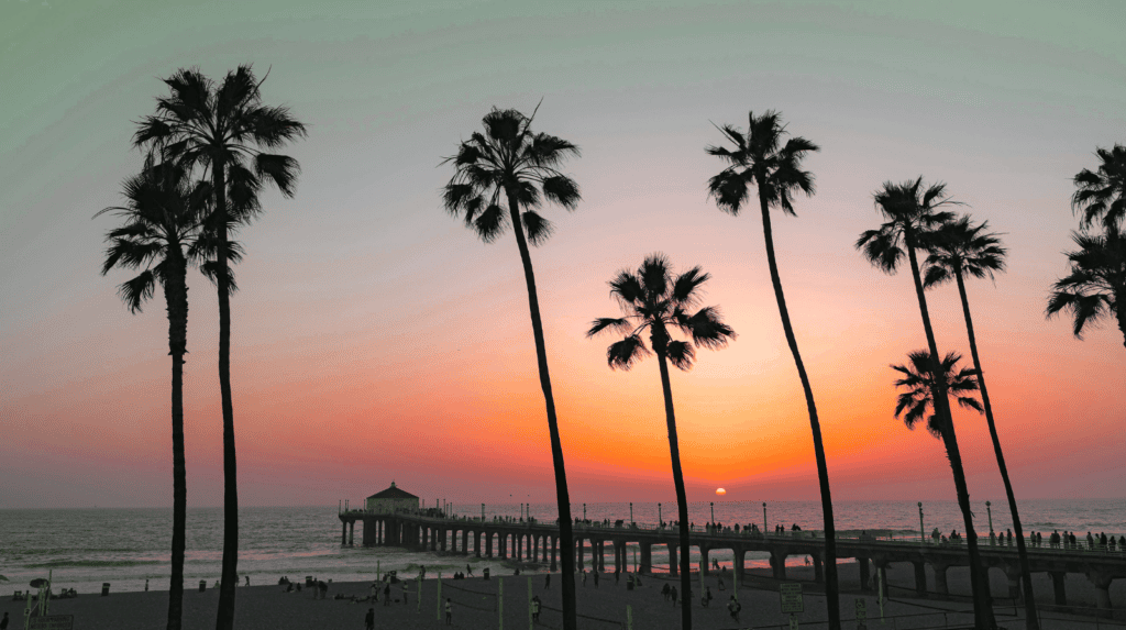 Tall palm trees frame a beach pier at sunset. The sky is orange and pink, and the sun is low over the ocean as people walk along the pier and relax on the sandy shore.