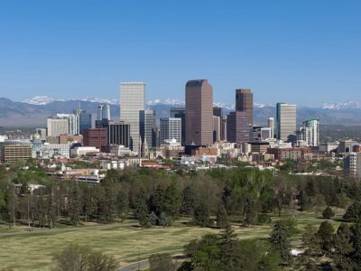 Denver skyline with tall buildings, green trees in the foreground, and snow-capped mountains in the background under a clear blue sky.