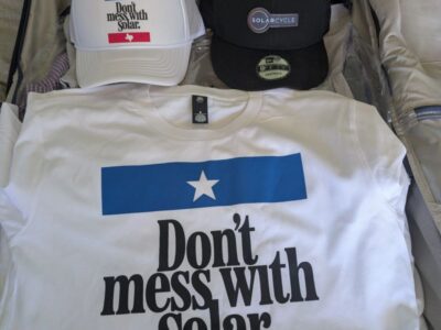 Open suitcase with two hats and a T-shirt inside. The white hat and T-shirt both read Dont mess with Solar. The black hat says SOLARCYCLE. The suitcase contains other folded clothes in the background.