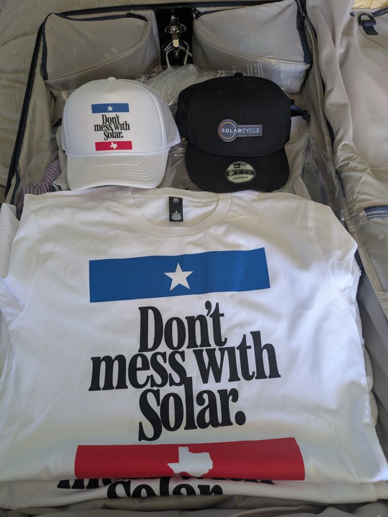 Open suitcase with two hats and a T-shirt inside. The white hat and T-shirt both read Dont mess with Solar. The black hat says SOLARCYCLE. The suitcase contains other folded clothes in the background.