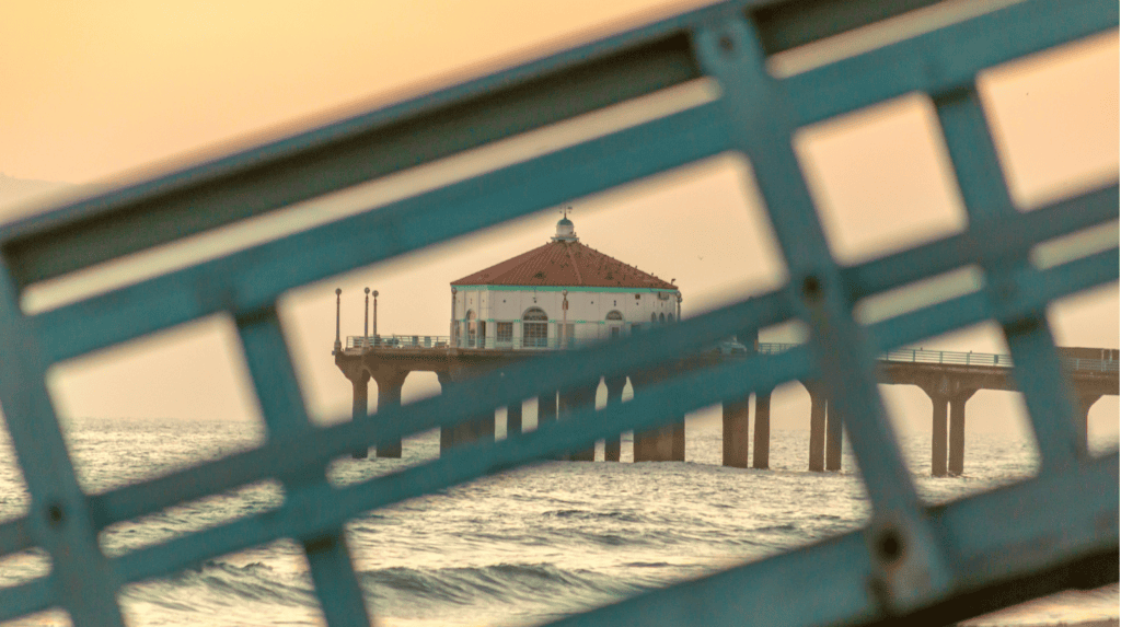 A building on a pier extends over the ocean at sunset, viewed through the diagonal bars of a nearby blue railing in the foreground.