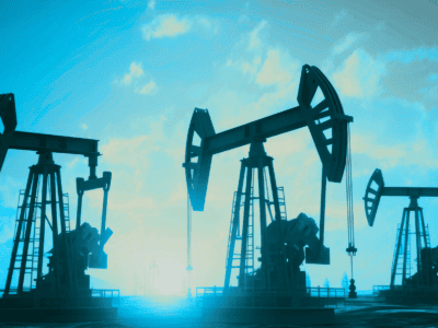 Three oil pumpjacks operate in a row on a barren landscape under a blue sky with scattered clouds, silhouetted by the setting or rising sun in the background.