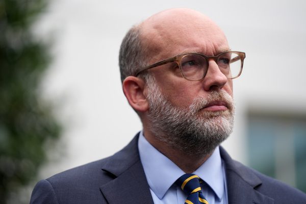 A man with glasses, a gray beard, and a receding hairline wears a suit, blue shirt, and striped tie, standing outdoors with a blurred building and greenery in the background.
