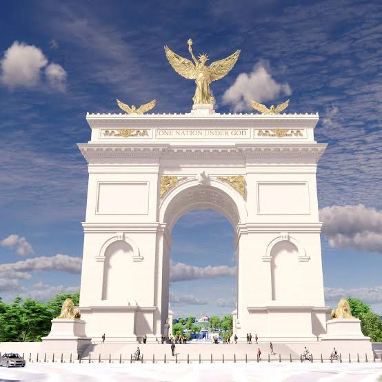 A large white triumphal arch with gold statues and One Nation Under God inscribed at the top, set against a blue sky with clouds and surrounded by greenery.