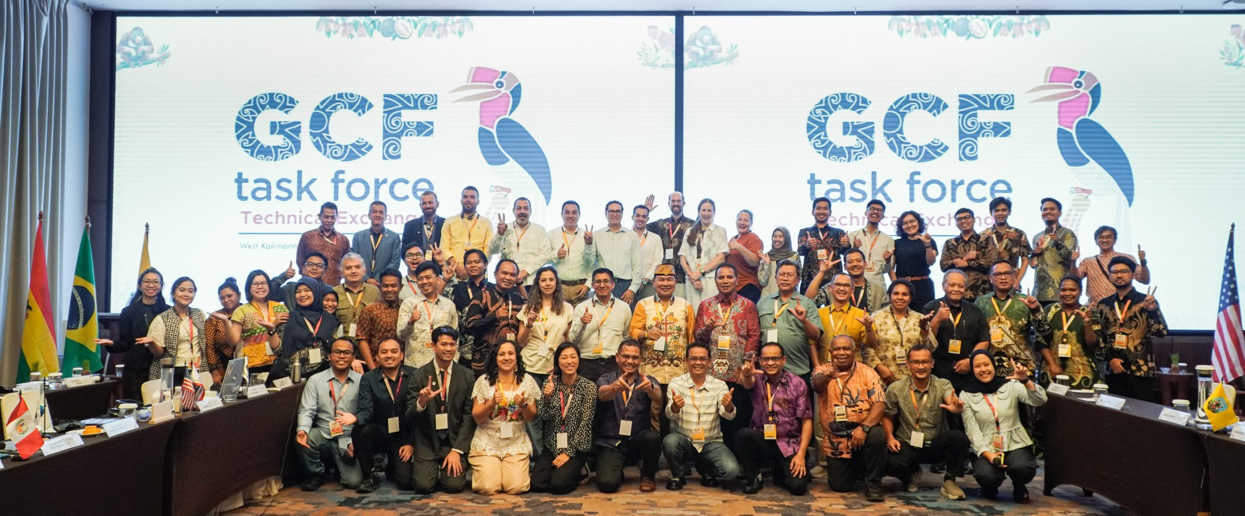 A large group of people pose for a photo in front of a screen displaying GCF task force Technical Exchange with a toucan logo and various national flags on the table in front of them.