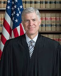 A man in a black judicial robe stands in front of an American flag and shelves of law books, smiling at the camera.
