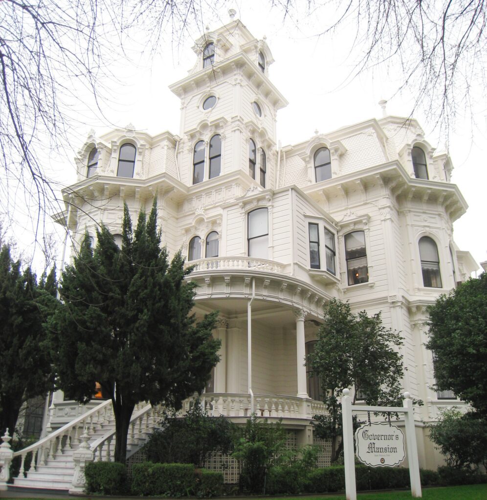 A large, ornate white Victorian mansion with a central tower, surrounded by greenery. A sign in front reads Governors Mansion..