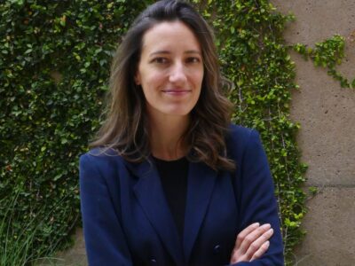 A woman with long brown hair, wearing a navy blazer, stands with arms crossed and smiles. She is outdoors in front of a wall covered with green climbing plants.