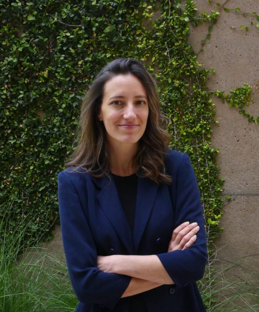 A woman with long brown hair, wearing a navy blazer, stands with arms crossed and smiles. She is outdoors in front of a wall covered with green climbing plants.