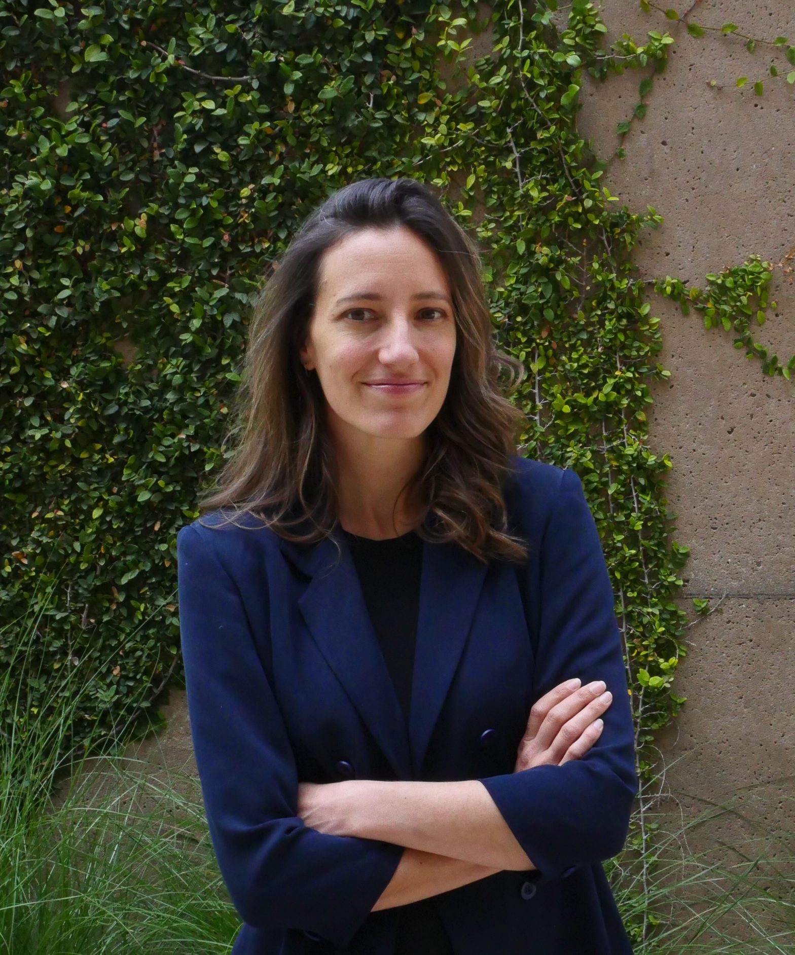 A woman with long brown hair, wearing a navy blazer, stands with arms crossed and smiles. She is outdoors in front of a wall covered with green climbing plants.