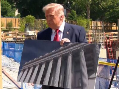 A man in a suit and red tie holds a large photograph of classical columns at a construction site with unfinished structures and trees in the background.