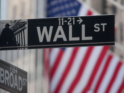 A Wall Street sign is in focus with a blurred American flag in the background. A Broad Street sign is partially visible below the Wall Street sign.