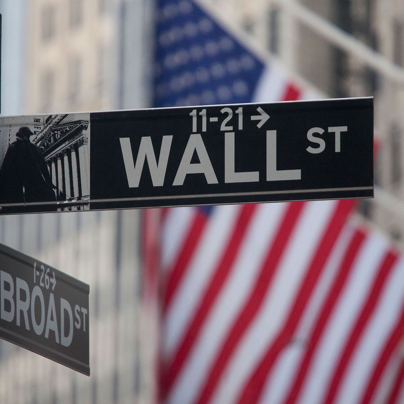 A Wall Street sign is in focus with a blurred American flag in the background. A Broad Street sign is partially visible below the Wall Street sign.