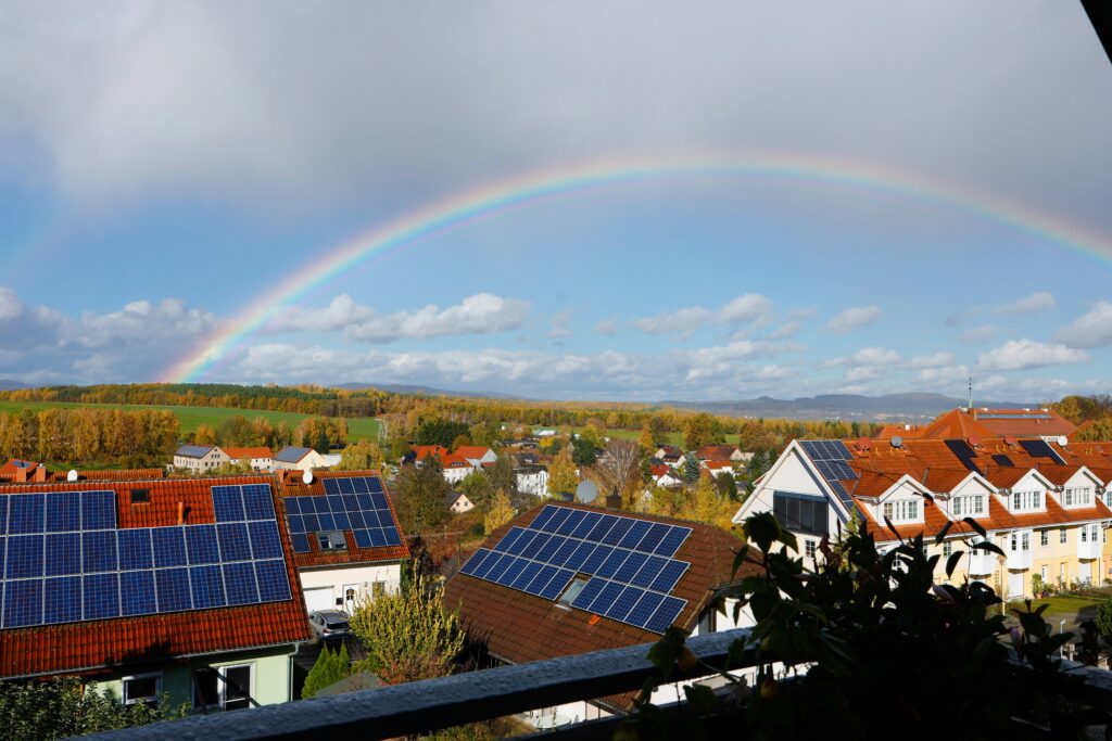 A rainbow arcs across a blue sky over a suburban neighborhood with red-roofed houses, many covered in solar panels, and green fields in the distance.