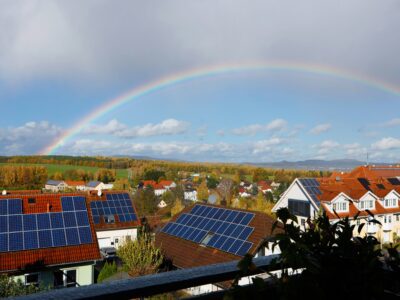 A rainbow arcs across a blue sky over a suburban neighborhood with red-roofed houses, many covered in solar panels, and green fields in the distance.