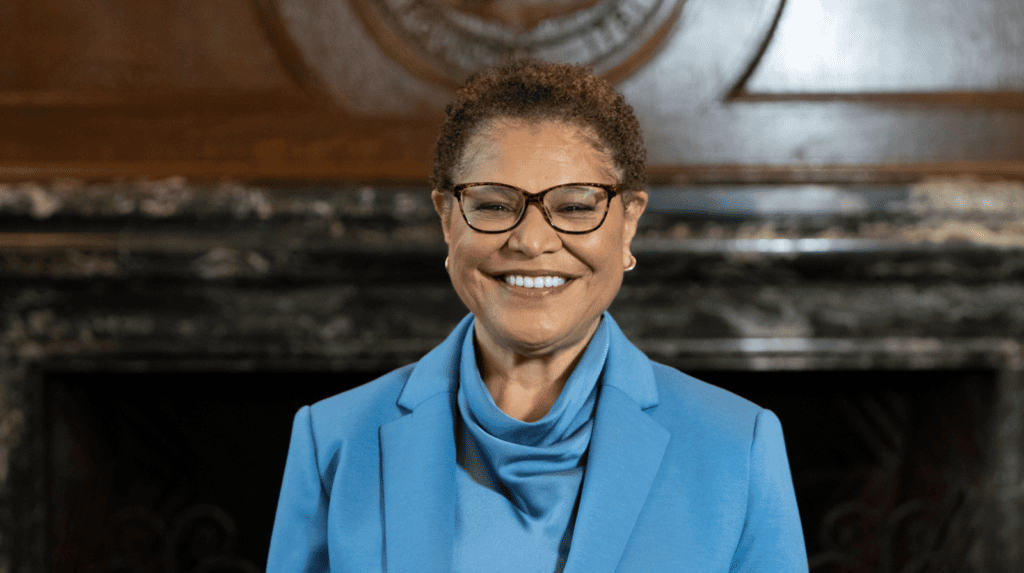 Karen Bass wearing a blue blazer and glasses, smiling in front of a fireplace.
