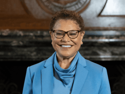 Karen Bass wearing a blue blazer and glasses, smiling in front of a fireplace.
