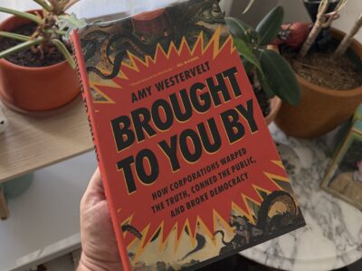 A hand holding the book Brought to You By by Amy Westervelt, featuring a red cover with yellow jagged graphics and illustration of octopus tentacles.