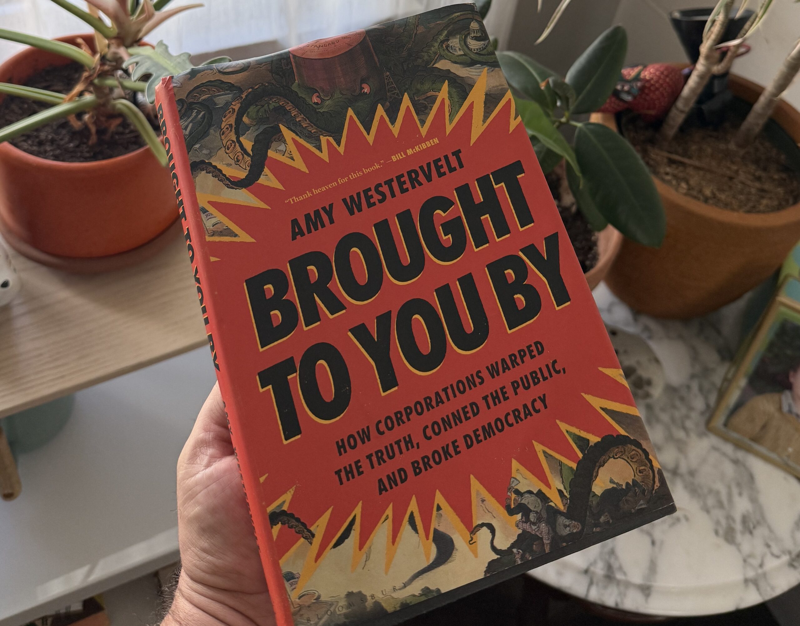 A hand holding the book Brought to You By by Amy Westervelt, featuring a red cover with yellow jagged graphics and illustration of octopus tentacles.