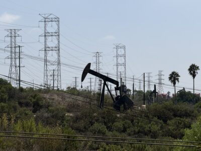 An oil pump jack operates in a landscape filled with large metal electricity transmission towers and low-lying shrubs.
