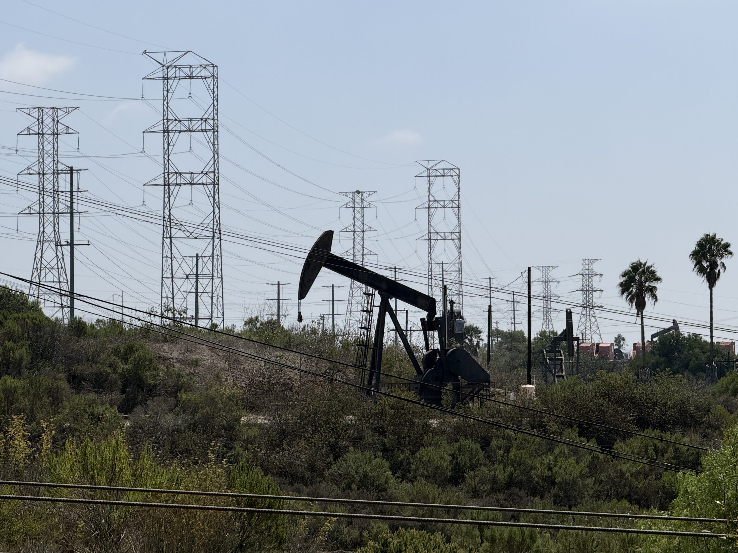 An oil pump jack operates in a landscape filled with large metal electricity transmission towers and low-lying shrubs.